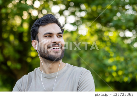 Close-up photo of a young smiling Indian man doing sports, standing in the park wearing headphones, smiling and looking to the side. Close-up photo of a young smiling Indian man doing sports, standing in the park wearing headphones, smiling and looking to the side. 125723365