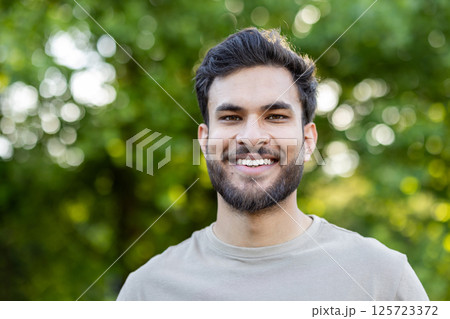 Smiling man with beard enjoying outdoors in park. Casual and relaxed expression conveys happiness and positivity. Sunlight filtering through leaves adds natural ambiance to portrait. 125723372
