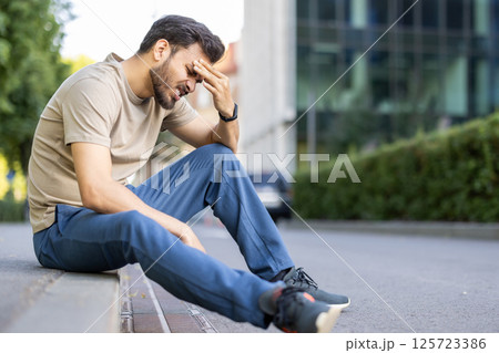 Tired and upset young muslim man sitting on the curb on the street and holding his head with his hand, thinking about problems. Tired and upset young muslim man sitting on the curb on the street and holding his head with his hand, thinking about problems. 125723386