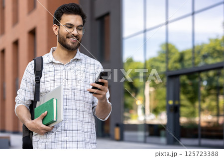 Smiling young Indian male student walking outside campus, holding books, backpack and using mobile phone. Smiling young Indian male student walking outside campus, holding books, backpack and using mobile phone. 125723388
