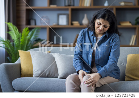 A young Muslim woman sits on the couch at home and holds her leg with her hands, feeling severe pain in her knee and bones. 125723429