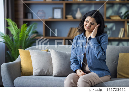 Muslim young woman in shirt sitting on sofa at home, grimacing in pain, holding hand to head, suffering from pressure. 125723432