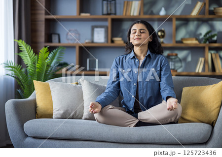 A young woman meditates on a grey sofa in her living room, promoting wellness and mindfulness. Interior home background. A young woman meditates on a grey sofa in her living room, promoting wellness and mindfulness. Interior home background. 125723436