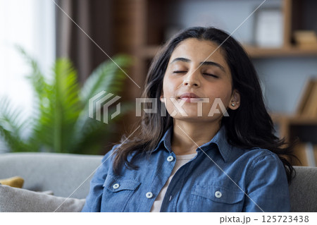 Young woman relaxing at home on the couch, eyes closed, practicing meditation and mindfulness for stress relief in a cozy living room. Young woman relaxing at home on the couch, eyes closed, practicing meditation and mindfulness for stress relief in a cozy living room. 125723438