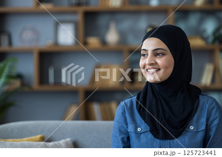 Close-up photo of a young Muslim woman wearing a black hijab sitting on a sofa at home and looking, to the side with a smile. Close-up photo of a young Muslim woman wearing a black hijab sitting on a sofa at home and looking, to the side with a smile. 125723441