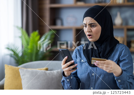 Worried middle eastern woman in hijab having problems with online payment using her credit card and her phone at home. 125723443