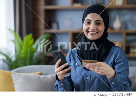 Portrait of a young Islamic woman in a hijab sitting on the sofa at home, holding a credit card and a mobile phone, looking at the camera with a smile. 125723444