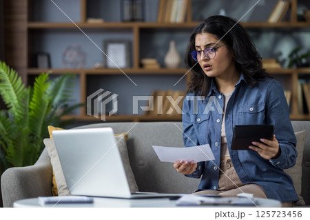 Worried young Muslim woman sitting at home on the couch behind a laptop, looking at the screen in disbelief and holding documents with a calculator in her hands. Worried young Muslim woman sitting at home on the couch behind a laptop, looking at the screen in disbelief and holding documents with a calculator in her hands. 125723456