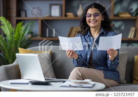 A smiling woman sitting on a sofa at home, holding documents and working with a laptop on the table. A smiling woman sitting on a sofa at home, holding documents and working with a laptop on the table. 125723457