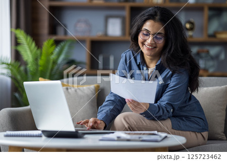 Smiling young Indian woman in casual clothes sitting on the couch at home and working remotely on a laptop, checking documents and bills. 125723462