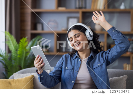 Happy young Indian woman sitting on the couch at home wearing headphones, holding a tablet, dancing and relaxing to music. Happy young Indian woman sitting on the couch at home wearing headphones, holding a tablet, dancing and relaxing to music. 125723472