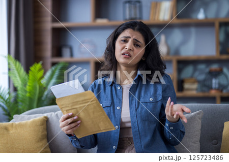 A young woman looks upset as she reads a document that she has just pulled from a brown envelope while at home on the couch. 125723486