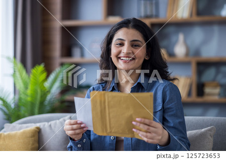 A young woman with a beaming smile sits on her couch, holding an envelope and letter, seemingly delighted by its contents. 125723653