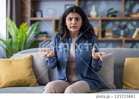 Upset Indian woman sitting on a sofa with a concerned expression. She is gesturing with her hands while looking directly at the camera. Upset Indian woman sitting on a sofa with a concerned expression. She is gesturing with her hands while looking directly at the camera. 125723661