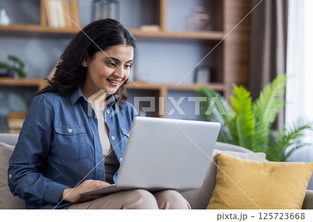 A young woman in a denim shirt smiles while using her laptop on the sofa at home, staying connected and productive. A young woman in a denim shirt smiles while using her laptop on the sofa at home, staying connected and productive. 125723668