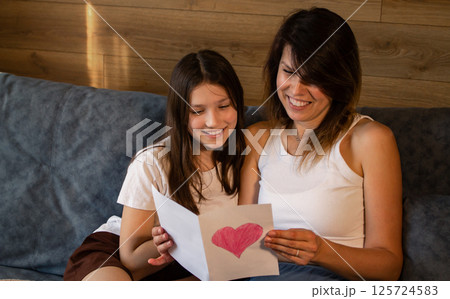 Happy mother's day concept. Mom and daughter sitting together on sofa in living room and reading greeting card. 125724583