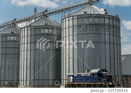 Large metal silos stand tall against the sky, storing harvested grain while a train is parked nearby at the agricultural site Large metal silos stand tall against the sky, storing harvested grain while a train is parked nearby at the agricultural site 125726418