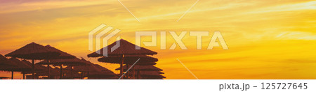 Tropical beach scenery with sun parasols. Straw sun umbrellas on the beach at purple-orange sunset. Gradient color. Horizontal banner Tropical beach scenery with sun parasols. Straw sun umbrellas on the beach at purple-orange sunset. Gradient color. Horizontal banner 125727645