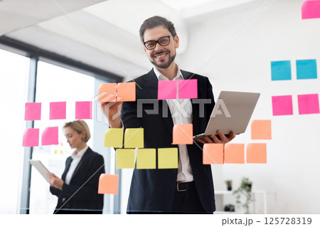 Businesswoman and businessman collaborating while using transparent glass board with sticky notes in modern office setting. Both professionals are organized and focused on task for effective project Businesswoman and businessman collaborating while using transparent glass board with sticky notes in modern office setting. Both professionals are organized and focused on task for effective project 125728319