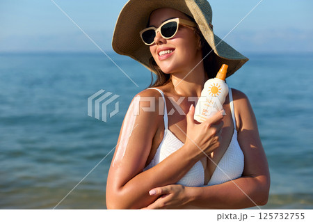 Woman applying sunscreen on her arm at the beach, enjoying a sunny day while wearing a  hat and sunglasses, emphasizing the importance of skin protection. 125732755