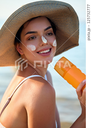 Beautiful young woman with sun cream on face. Girl holding sunscreen bottle on the beach and applying moisturizing lotion on skin.Skin care. Sun protection. Suntan Beautiful young woman with sun cream on face. Girl holding sunscreen bottle on the beach and applying moisturizing lotion on skin.Skin care. Sun protection. Suntan 125732777