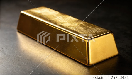 Close-up of a shiny gold bar with smooth surface and rectangular shape, resting on a reflective dark surface with soft lighting highlighting its metallic texture 125733426