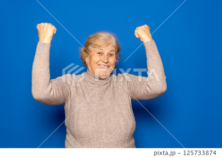 Caucasian woman in her 70s wearing a dark beige sweater celebrating happily with her fists raised, isolated on blue studio background. 125733874