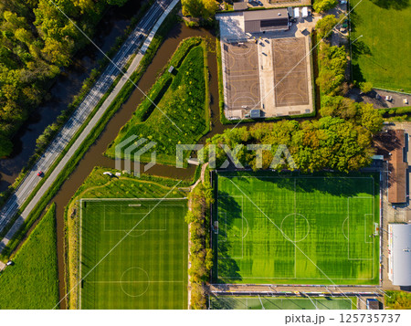 Aerial view from above of a green sports field with white markings, surrounded by trees and footpaths, with bright sunlight casting long shadows. A highway with cars driving on it. Aerial view from above of a green sports field with white markings, surrounded by trees and footpaths, with bright sunlight casting long shadows. A highway with cars driving on it. 125735737