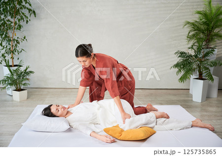 Masseuse making thai yoga massage. Woman therapist doing traditional massage treatment, stretching female patient's body. Client lying on mat, enjoying hand massage in calm atmosphere. Masseuse making thai yoga massage. Woman therapist doing traditional massage treatment, stretching female patient's body. Client lying on mat, enjoying hand massage in calm atmosphere. 125735865