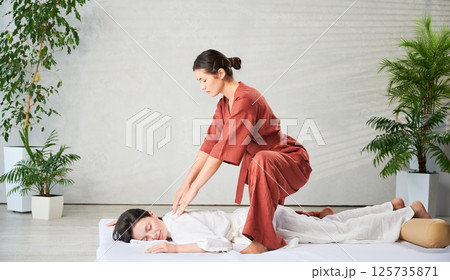 Masseuse making thai yoga massage. Woman therapist doing traditional massage treatment for female patient indoors. Client lying on mat, meditating, enjoying hand massage in calm atmosphere. Masseuse making thai yoga massage. Woman therapist doing traditional massage treatment for female patient indoors. Client lying on mat, meditating, enjoying hand massage in calm atmosphere. 125735871