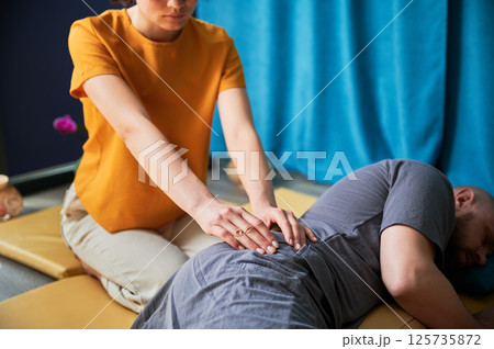 Masseuse making thai yoga massage. Cropped view of woman therapist doing traditional massage treatment for male patient indoors. Client lying on mat, enjoying hand massage in calm atmosphere. 125735872
