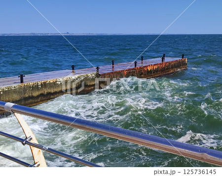 Stormy sea. Large sea waves crashing and hitting against old concrete pier on sunny summer day. High waves with white foam. Big waves breaking, spreading on cement pier. Splashes bursts and splashing Stormy sea. Large sea waves crashing and hitting against old concrete pier on sunny summer day. High waves with white foam. Big waves breaking, spreading on cement pier. Splashes bursts and splashing 125736145