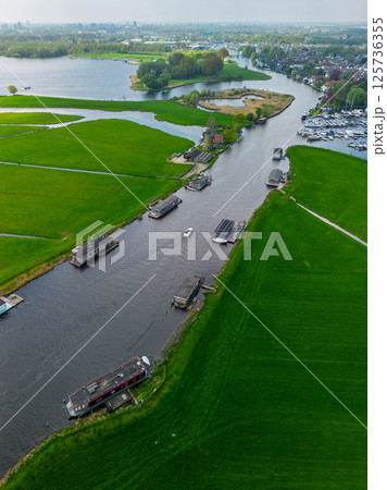Aerial view of a peaceful Dutch landscape with green fields, a canal lined with houseboats, a traditional windmill, and a marina in the distance under a cloudy sky. 125736355