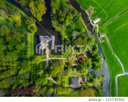 Aerial view of a historic castle surrounded by a forest, with vast green fields, winding waterways, and a river stretching across the flat Dutch countryside under a partly cloudy sky. Aerial view of a historic castle surrounded by a forest, with vast green fields, winding waterways, and a river stretching across the flat Dutch countryside under a partly cloudy sky. 125736362