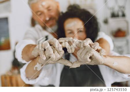 Mutual creative work. Adult elegant couple in casual clothes and aprons. People creating a bowl on a pottery wheel in a clay studio. 125743550
