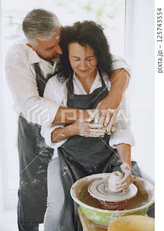 Mutual creative work. Adult elegant couple in casual clothes and aprons. People creating a bowl on a pottery wheel in a clay studio. 125743554