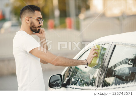 Young man in fashion clothes washing car at car wash station using washcloth. Man wearing white t-shirt and glasses and talking on the phone. Indian man washing his white vehicle. Young man in fashion clothes washing car at car wash station using washcloth. Man wearing white t-shirt and glasses and talking on the phone. Indian man washing his white vehicle. 125743571