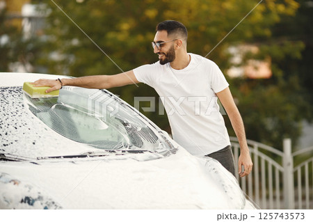 Young man in fashion clothes washing car at car wash station using washcloth. Man wearing white t-shirt and jeans. Indian man washing his white vehicle. Young man in fashion clothes washing car at car wash station using washcloth. Man wearing white t-shirt and jeans. Indian man washing his white vehicle. 125743573