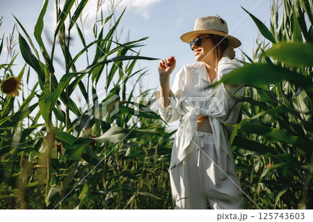 Pregnant woman walking in a maize field. Brunette woman walking in summer field wearing white clothes. Woman wearing sunglasses and straw hat. 125743603