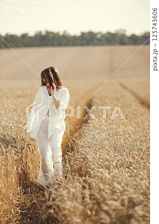 Woman running in wheat field. Brunette woman walking in summer field wearing white clothes. Woman posing for a photo. 125743606