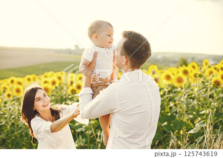 Photo of a young family at the sunflowers field on a sunny day. Brunette mother, father and their little blonde son posing for a photo. Man and woman playing with their son. 125743619