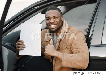 Black man holding blank sheet of paper in his hand. Man sitting in a car and wearing brown jacket. Man has happy face impression and thumb up. 125743635