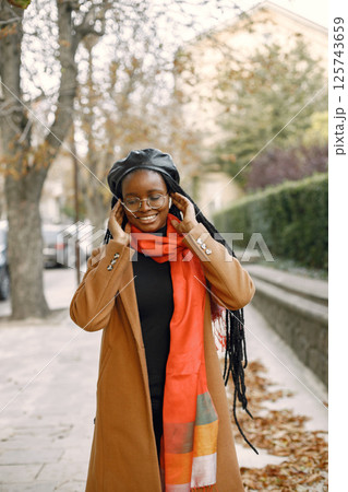 Young black woman with a long locs hairstyles standing outside. Woman wearing brown coat, orange scarf and black hat. Woman in glasses using a white earphones. 125743659