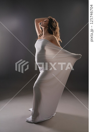 Beautiful pregnant woman posing gracefully in a flowing white garment Beautiful pregnant woman posing gracefully in a flowing white garment 125746704