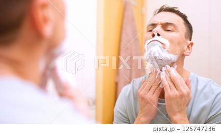 Man applying shaving cream on face in bathroom mirror 125747236