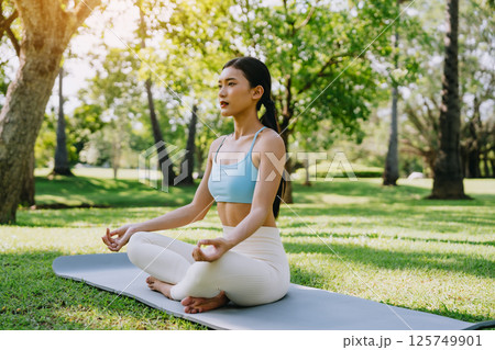 Asian woman meditating in a peaceful park setting, sitting cross-legged on green grass 125749901