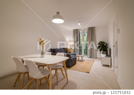 Minimalist dining and living area with white dining table, chairs, gray sofa, and window with natural light Minimalist dining and living area with white dining table, chairs, gray sofa, and window with natural light 125751832