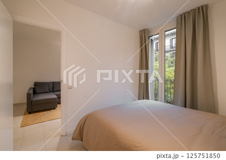 Bedroom interior with taupe bedspread, large window with beige curtains and view of living area Bedroom interior with taupe bedspread, large window with beige curtains and view of living area 125751850