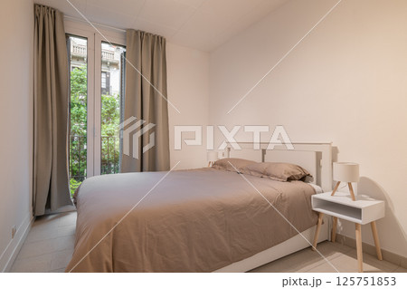 Simple bedroom interior with double bed, taupe bedding and large window with beige curtains Simple bedroom interior with double bed, taupe bedding and large window with beige curtains 125751853