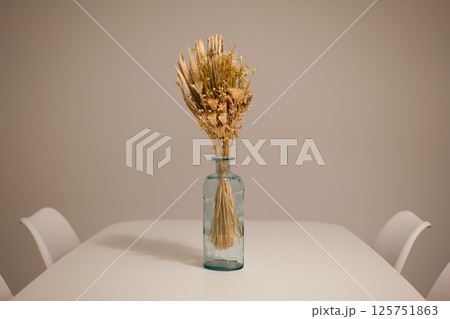 Dining table with white surface and blue glass vase holding dried flowers centered between white chairs 125751863